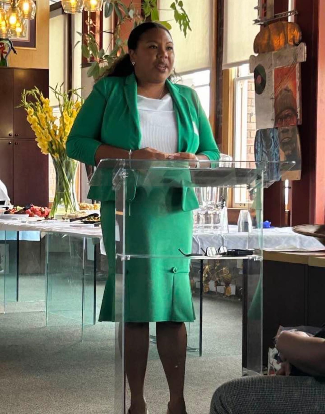 Woman in a green suit speaking at a clear podium in a bright room.