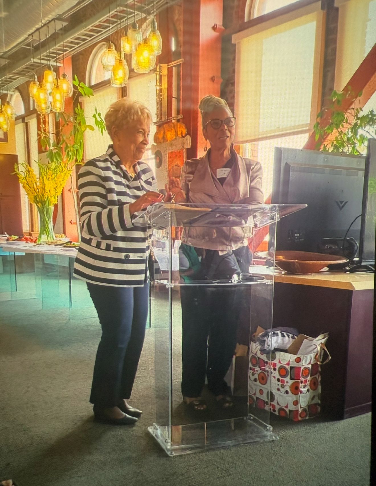 Two people stand at a clear podium in a warmly lit restaurant, speaking beside a monitor.