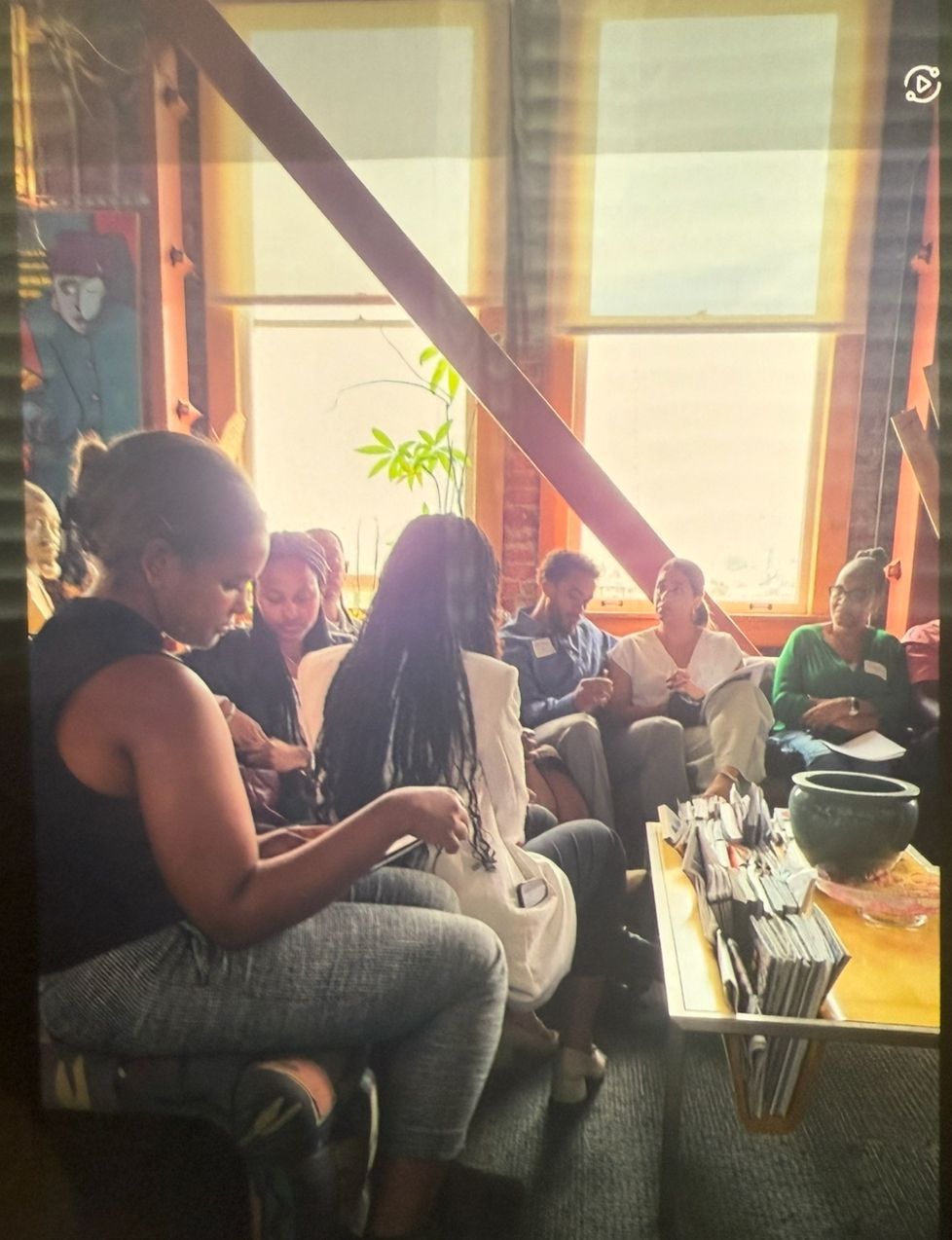 People seated in a sunlit living room, one person knitting while others chat on a couch.