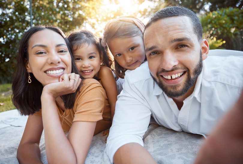 Four smiling people lying on a blanket outdoors, posing closely together in warm sunlight