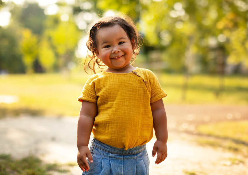 Smiling child in a yellow shirt and jeans standing outdoors on a sunny path.