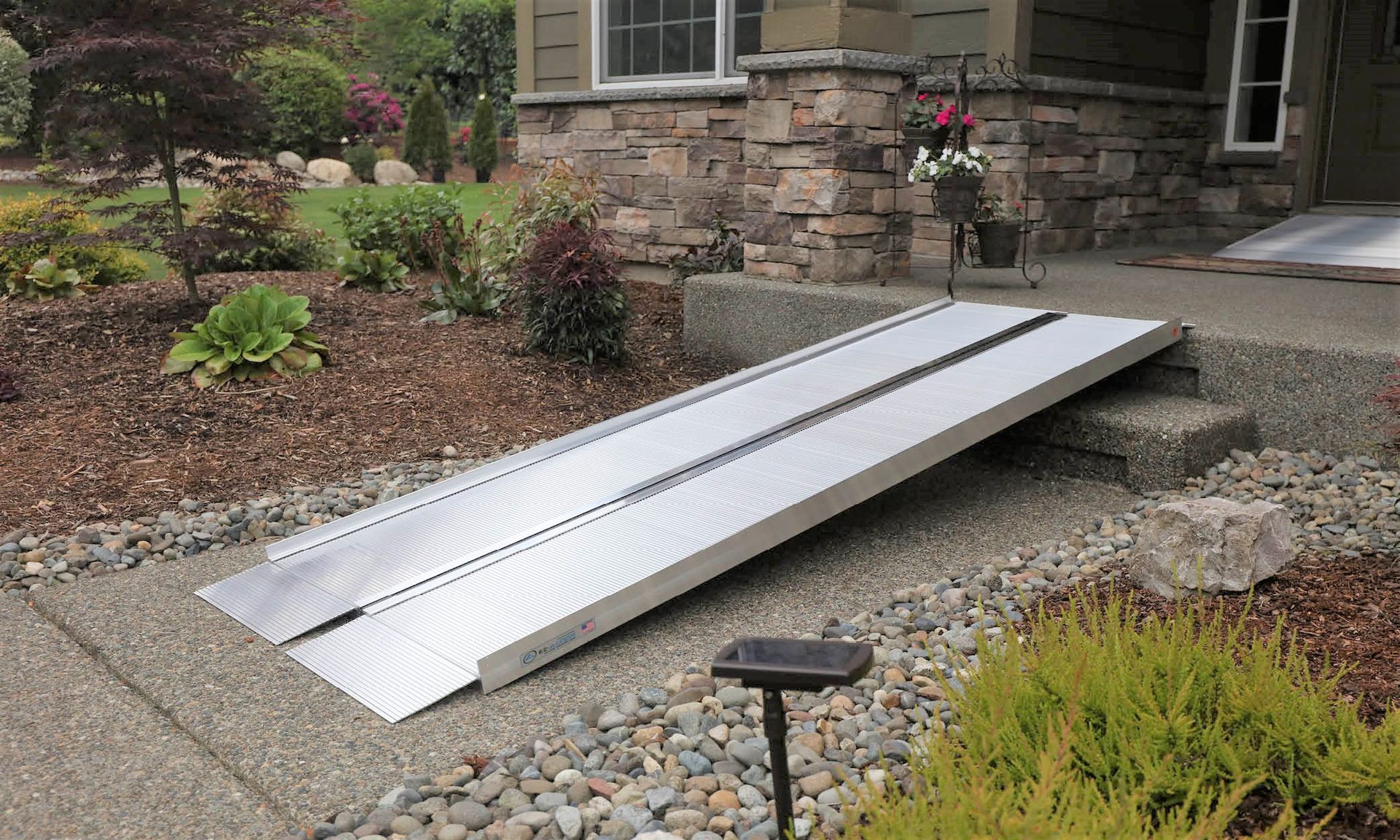 Silver wheelchair ramp leading to a stone house entrance, set in a gravel garden.