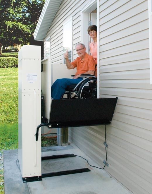 Man in a wheelchair using a home vertical platform lift beside a house, with a person standing behind him