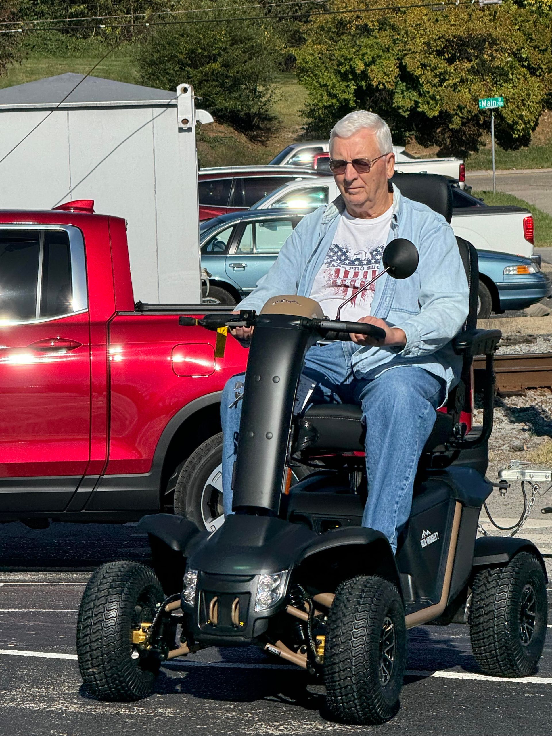 Man in a blue shirt driving a black mobility scooter beside a red pickup truck in a parking lot