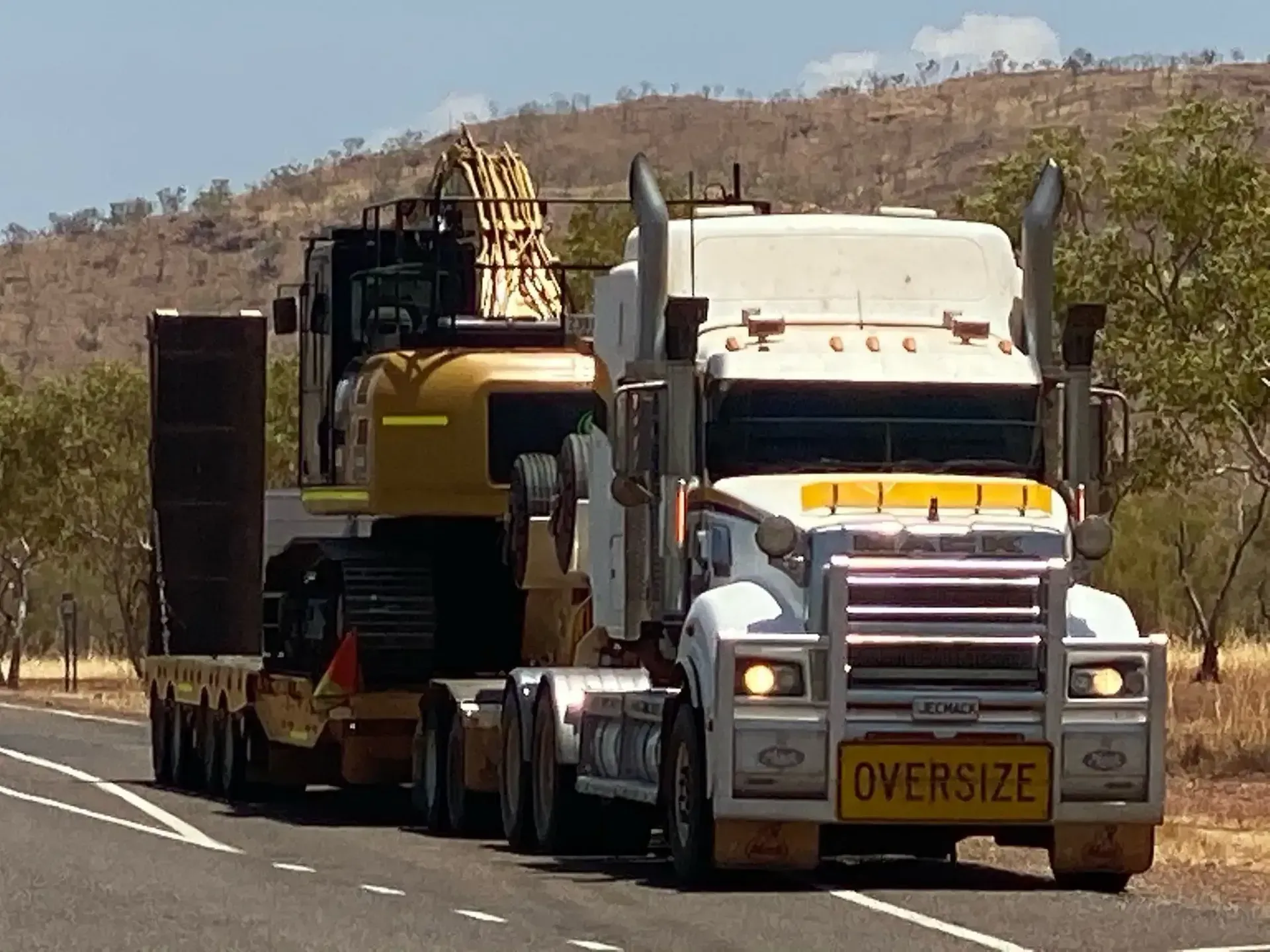 A White Truck is Driving Down a Street With Towing A Large Excavator — JEC Transport in Winnellie, NT