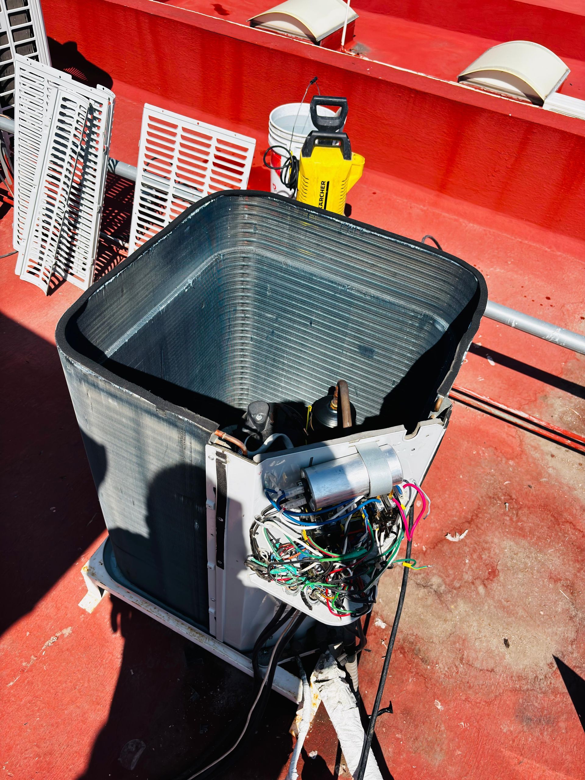 Una unidad exterior de aire acondicionado con la cubierta superior retirada, dejando al descubierto la bobina interna y el cableado, sobre un tejado rojo.