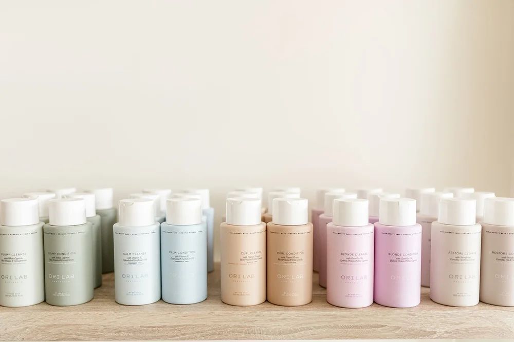 Pastel-coloured Skincare Bottles and Jars Lined Up on a Shelf Against a Beige Wall — Hair Truss & Co In Wallsend, NSW