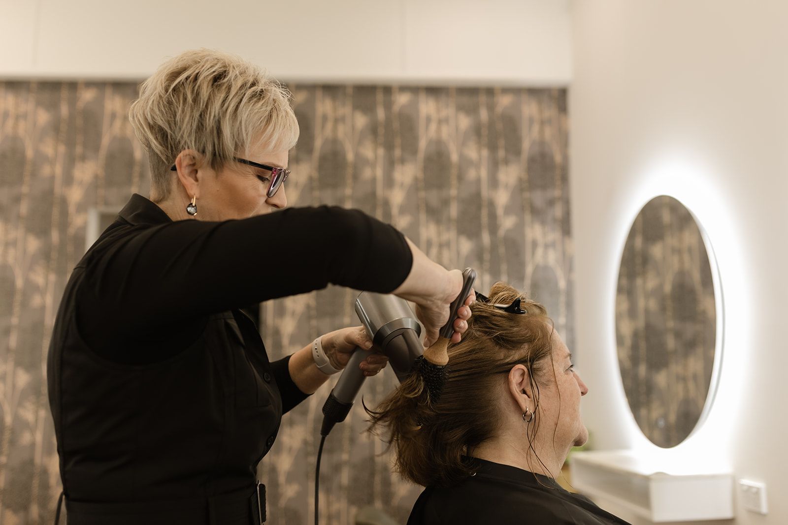 Hairdresser Styling a Seated Client’s Hair Near a Round Mirror in a Salon — Hair Truss & Co In Wallsend, NSW
