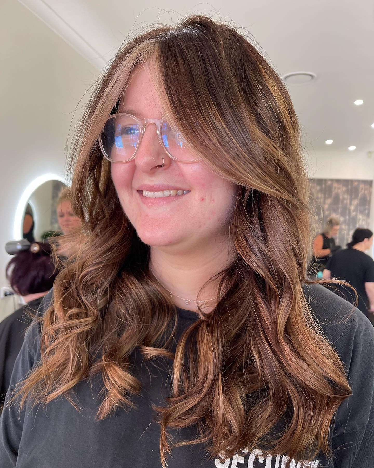 Person With Long Wavy Brown Hair and Glasses Smiling Indoors at a Social Event — Hair Truss & Co In Wallsend, NSW