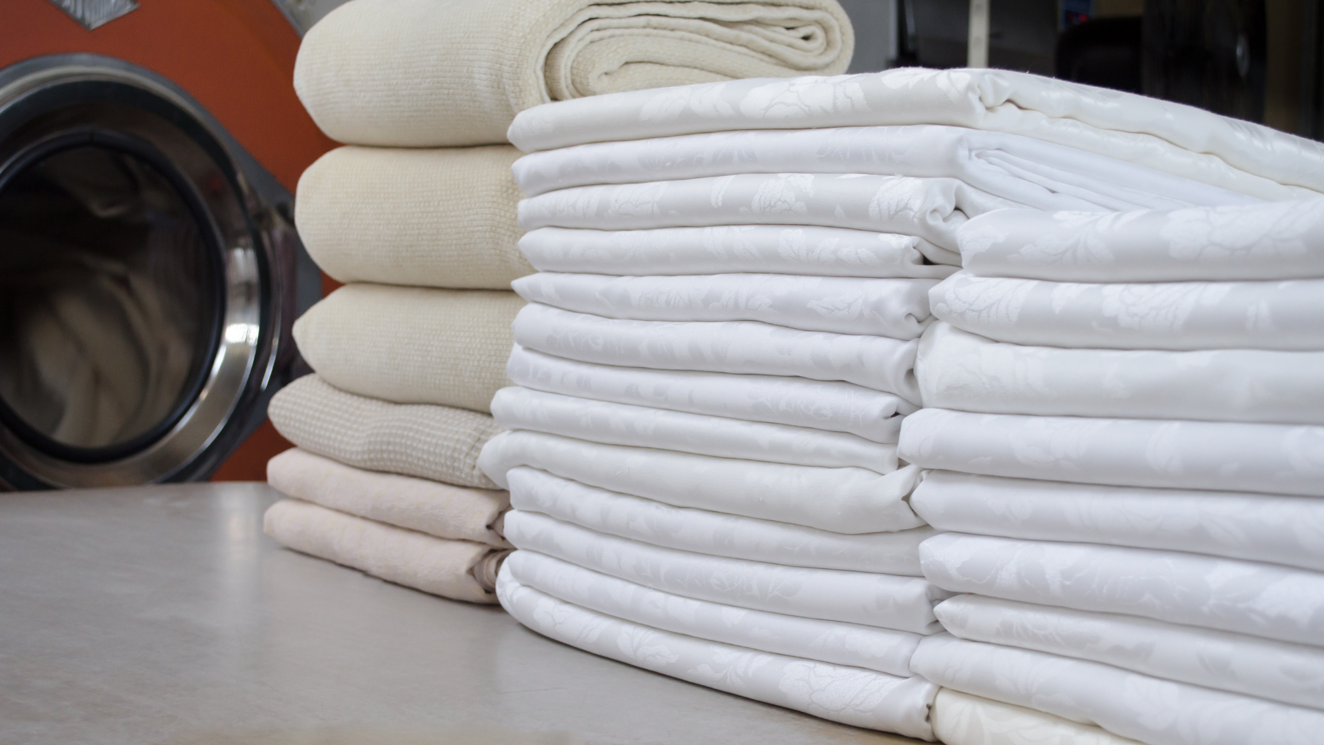 Stacks of folded white and beige linens on a counter with a commercial washing machine visible in the background.