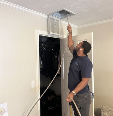 Technician standing on a ladder adjusting a ceiling air vent with a long tool in a hallway.