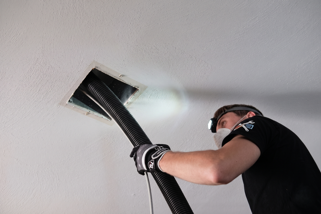 Man using a vacuum hose to clean a ceiling vent in a white room