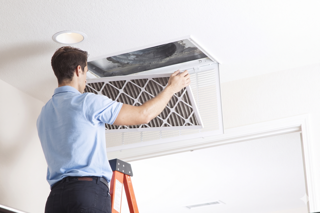 Worker cleaning a ceiling HVAC vent on a ladder in a bright room