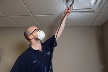 Worker in a mask reaching up to inspect a ceiling tile in a room