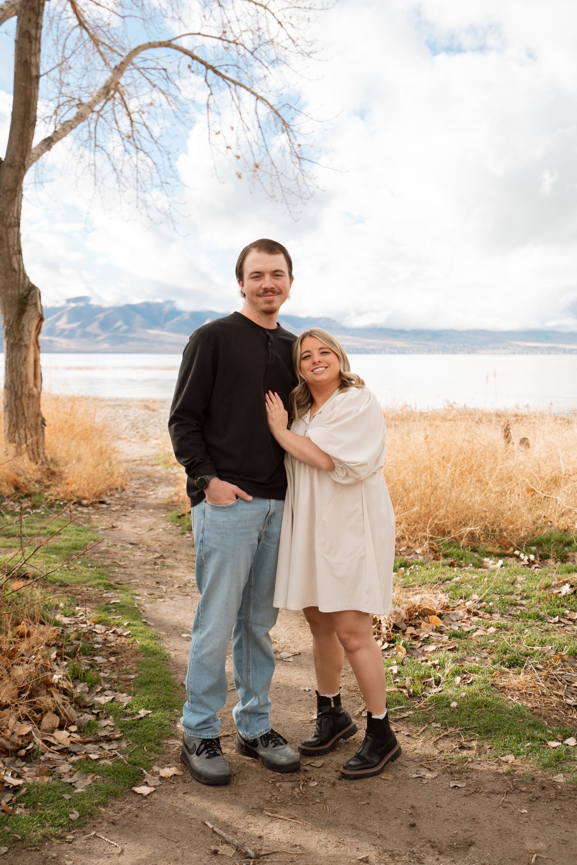 Couple standing by a lakeside, with autumn trees and mountains in the background.