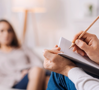 A man is writing in a notebook while a woman sits in the background.