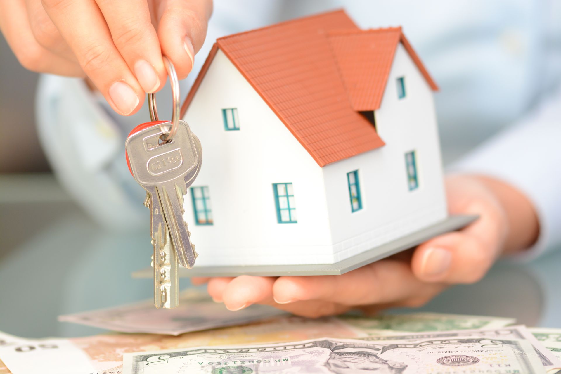Hand holding house key above a small model house and money