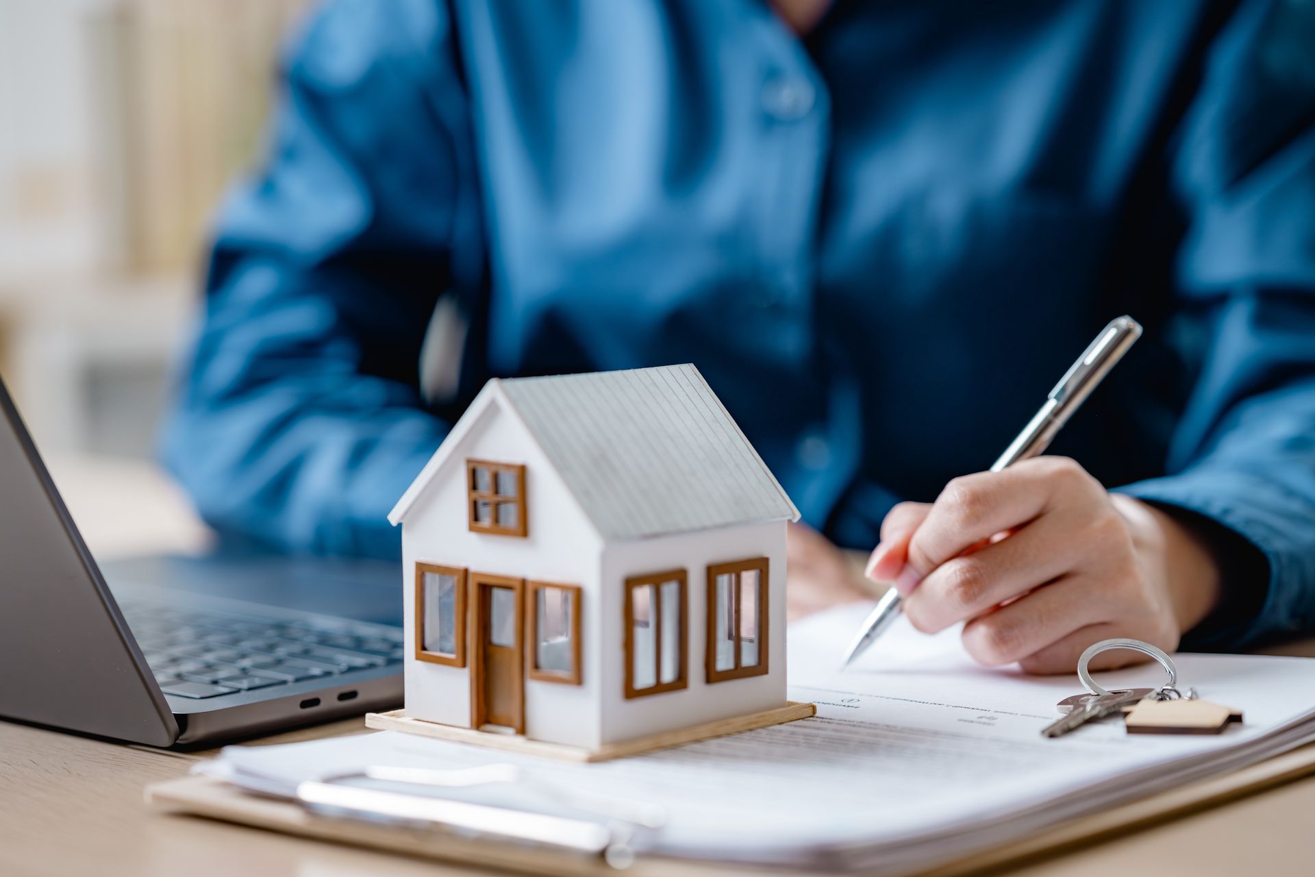 Person writing beside a small model house and laptop on a desk, suggesting home planning or real estate work