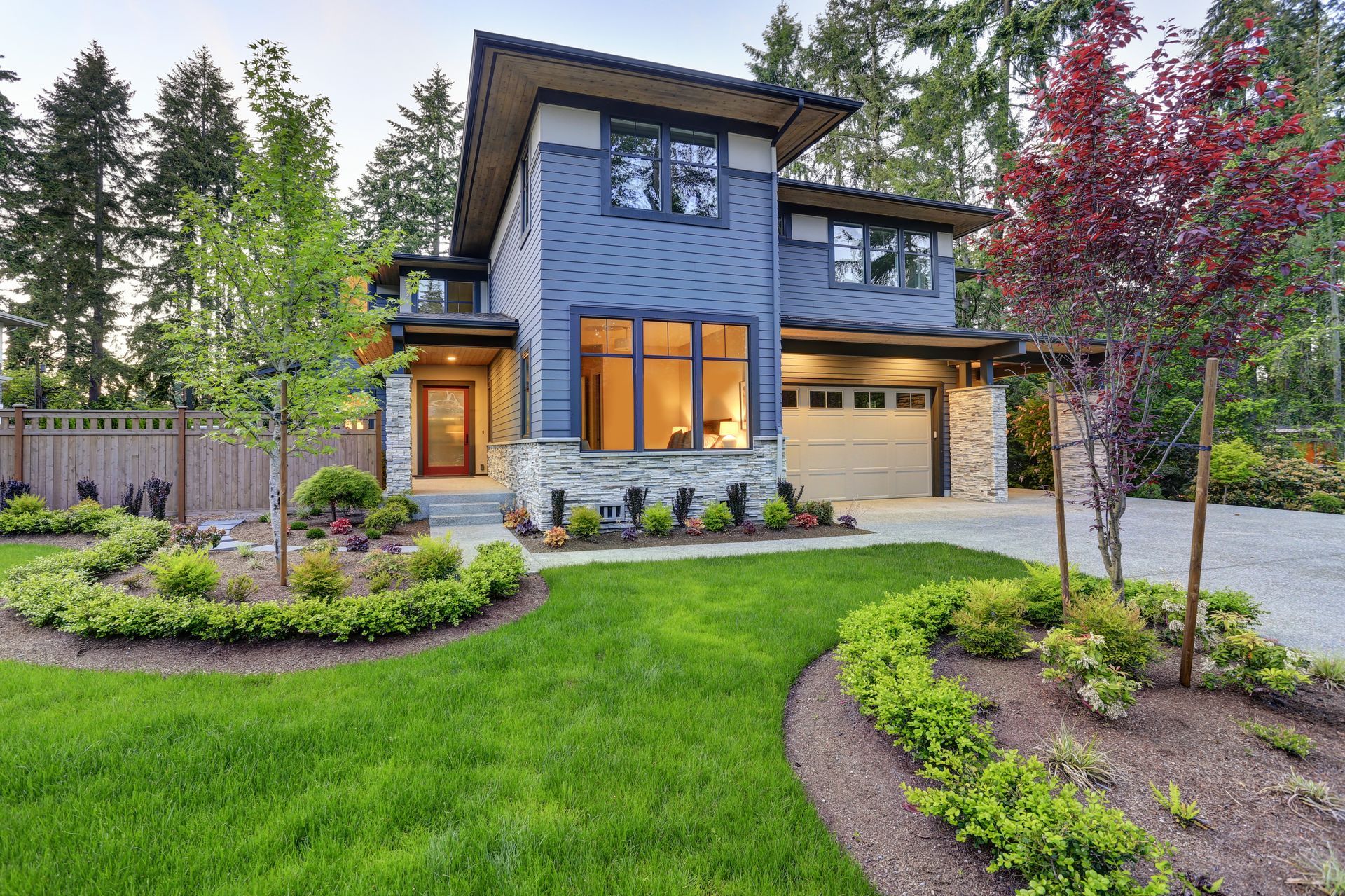 Modern two-story blue house with landscaped front yard and glowing porch lights at dusk