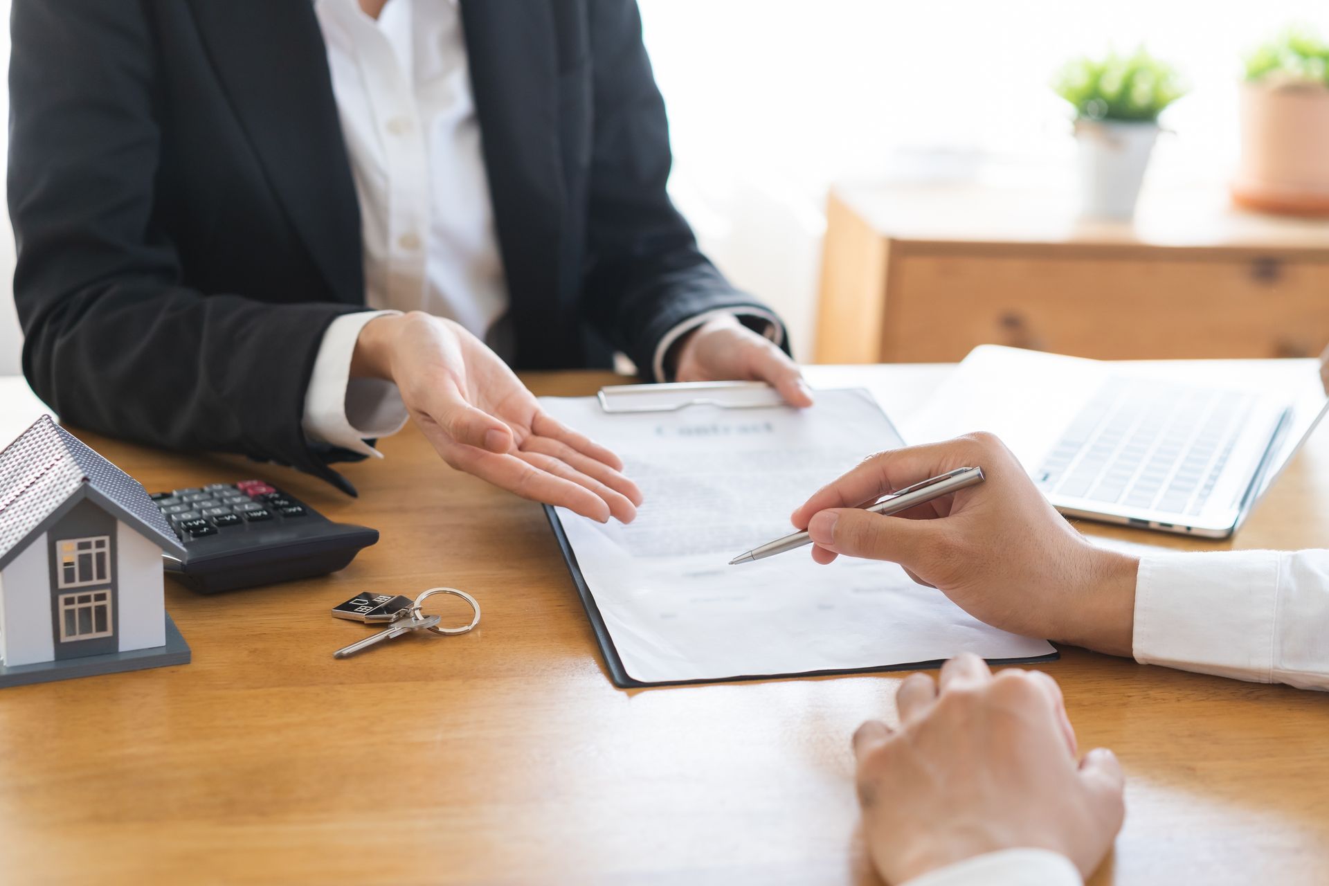 Business meeting with two people signing documents at a wooden table, with a house model and keys nearby. Business meeting with two people signing documents at a wooden table, with a house model and keys nearby.