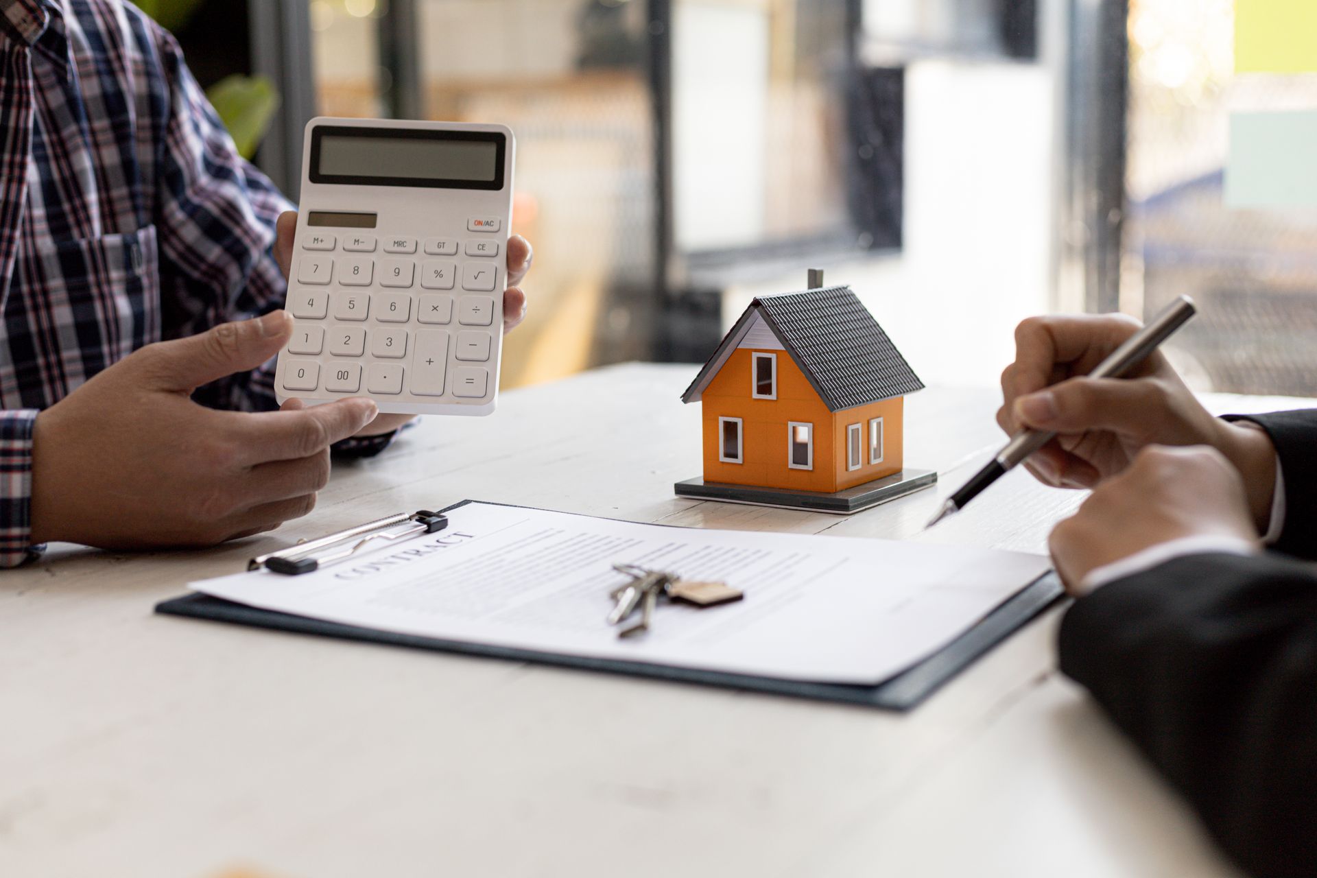 Hands using calculator and pen beside a small house model on papers at a desk