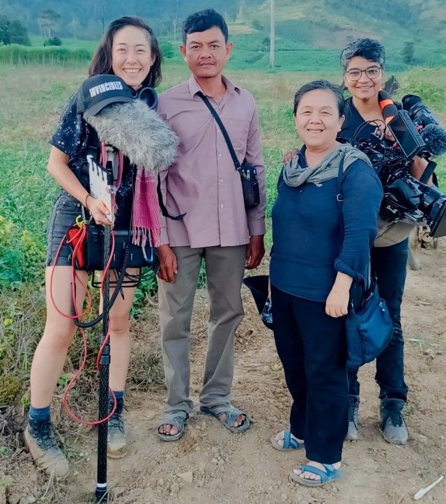 Four people, two with cameras, stand outdoors. A woman with sound equipment smiles. Others look at the camera, smiling.