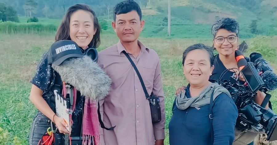 Four people smiling for a photo in a field. One holds a microphone, another, a camera.