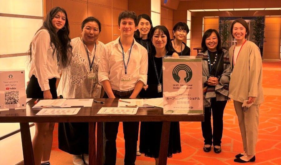 Group of people at a table with a sign, possibly at a conference. Smiling, diverse, indoors with orange carpet.