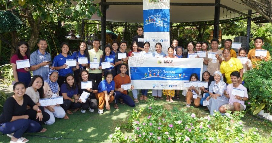 A large group of people posing for a photo outdoors while holding certificates, with a banner in the center.