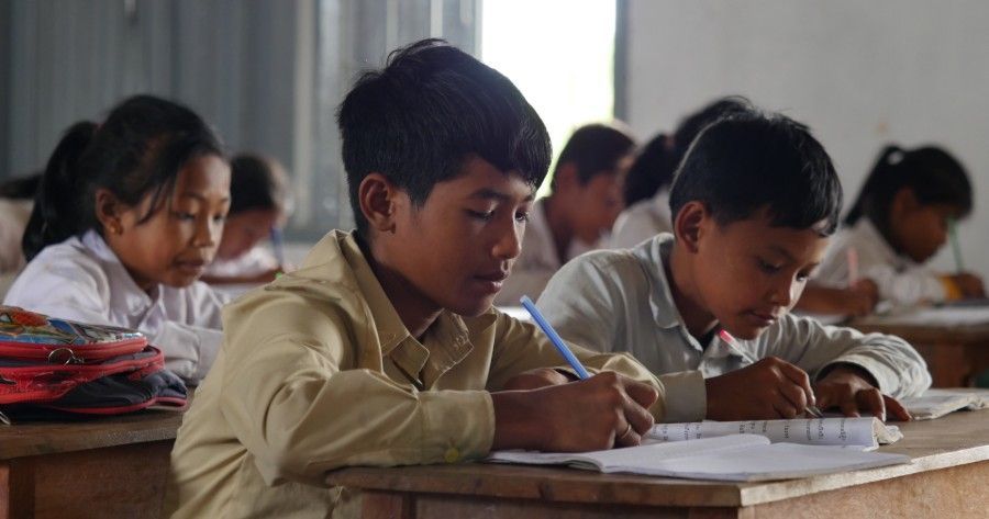 Students writing in a classroom with notebooks and pencils at shared desks