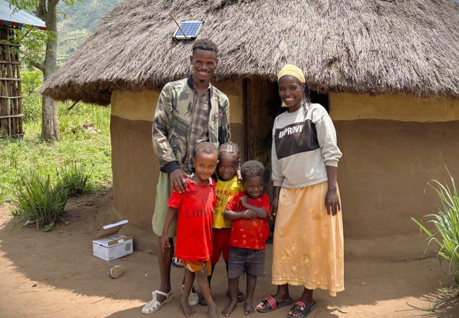 Family standing outside a thatched hut, with two children in front and a small solar panel on the roof