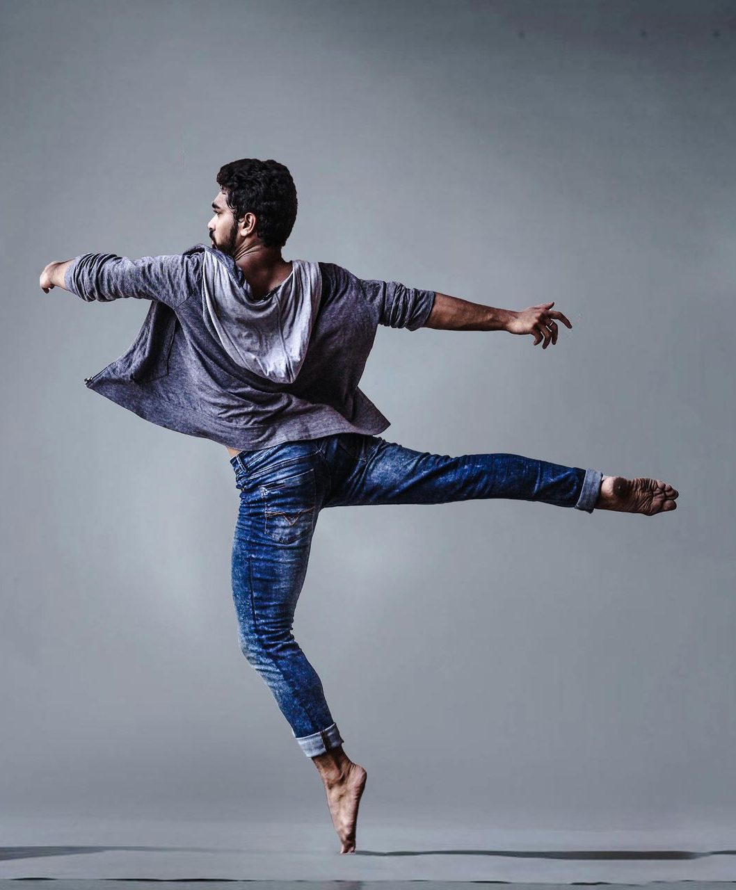 A person in a loose hoodie and blue jeans is captured in dance balance on tippy toes of one foot