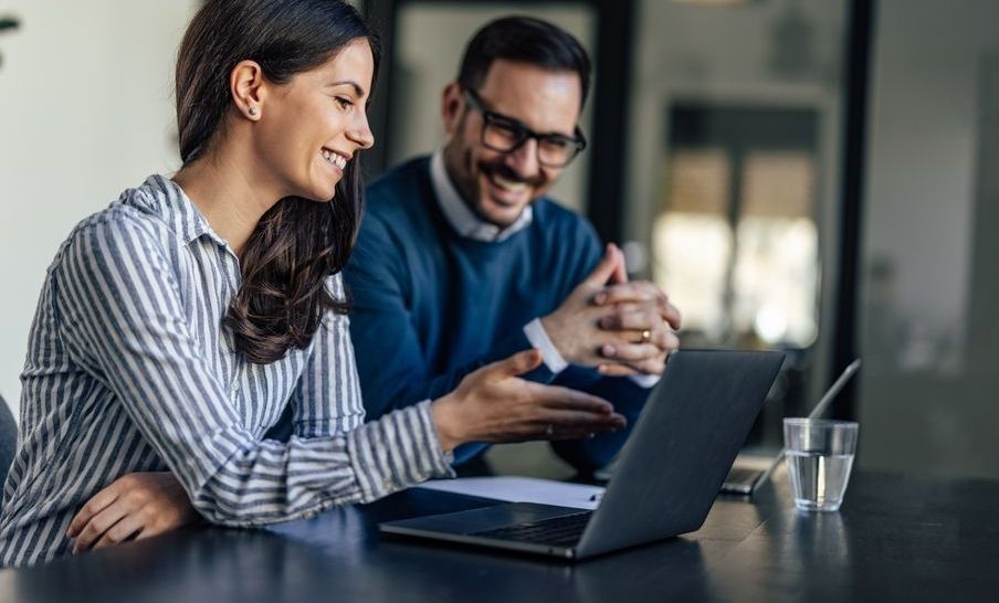 A man and a woman are sitting at a table looking at a laptop computer.