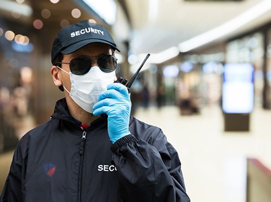 A security guard wearing a mask and gloves is talking on a walkie talkie.