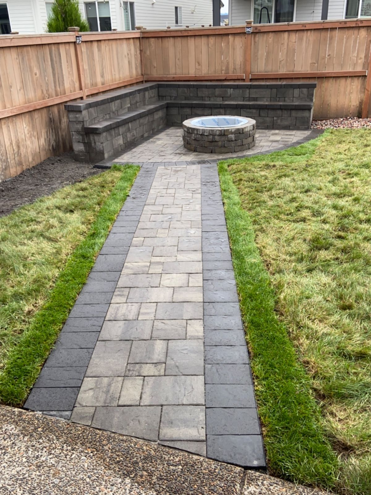 A brick walkway leading to a fire pit in a backyard.