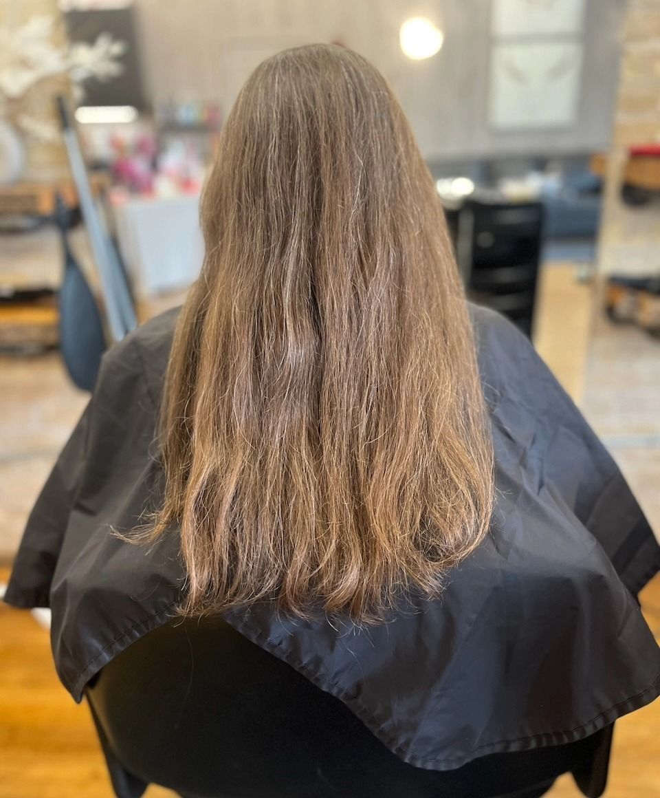 Back view of a person with long brown hair in a salon chair, wearing a black cape.