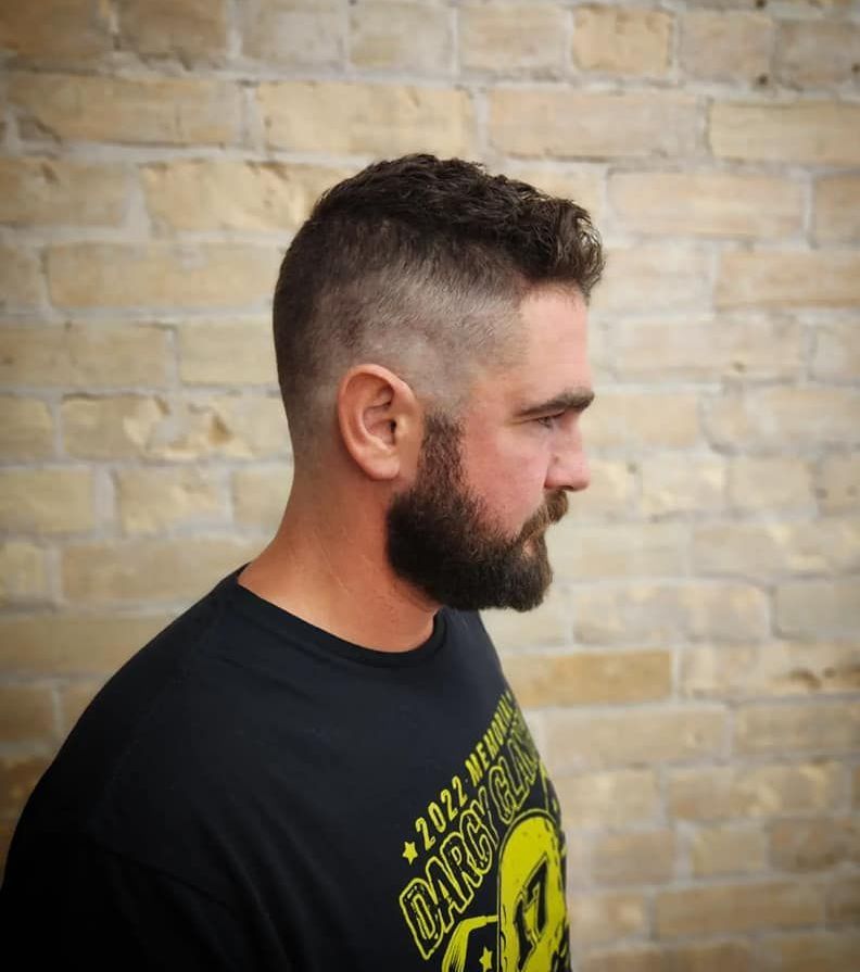 Side-profile portrait of a bearded man with a short fade haircut against a tan brick wall.