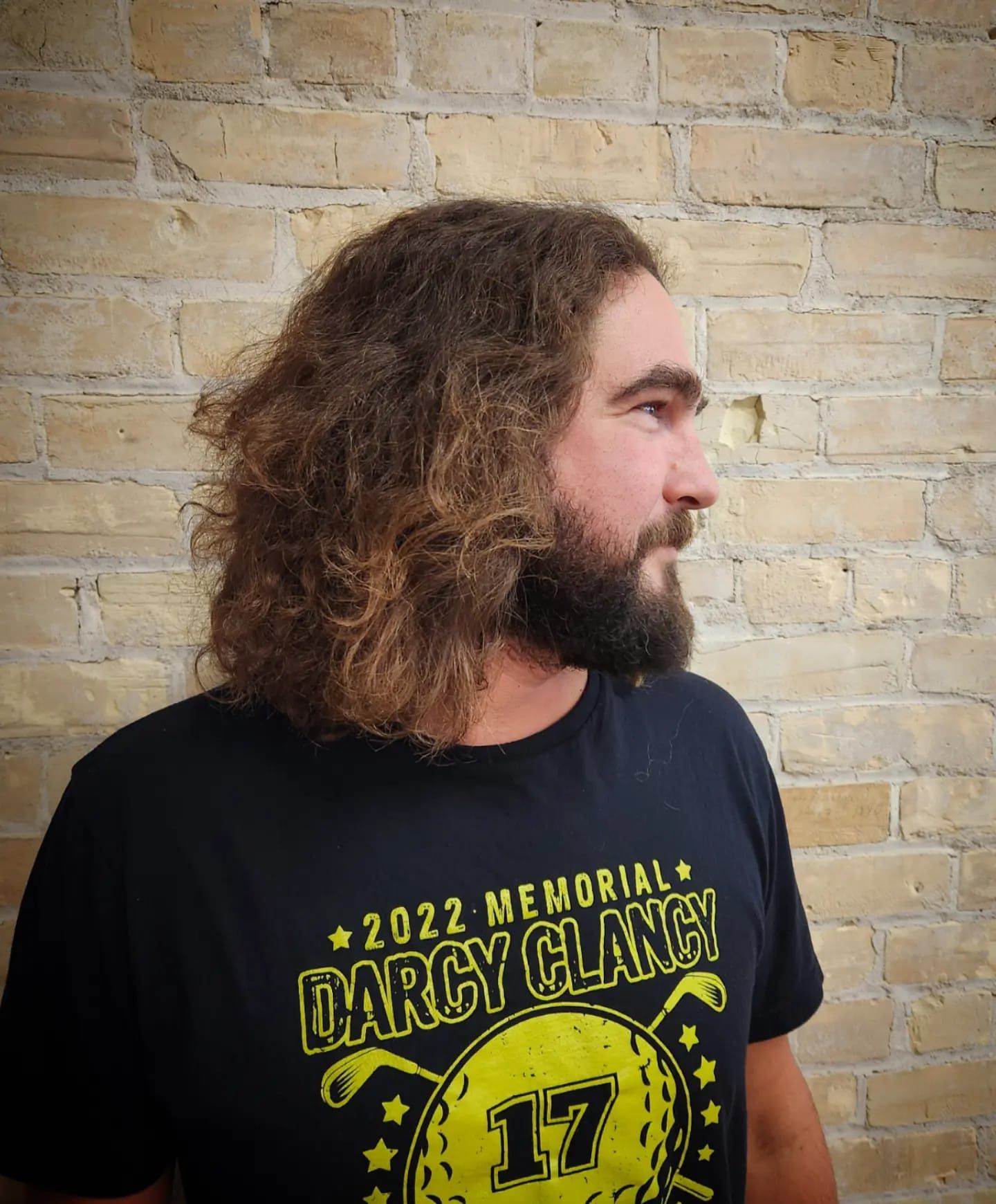 Profile view of a bearded person with curly hair in a black T-shirt against a brick wall.