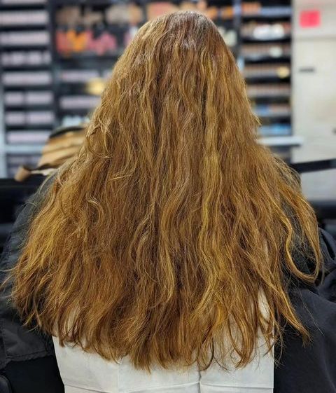 Person with long wavy light brown hair seen from behind in a salon chair, facing a mirror.