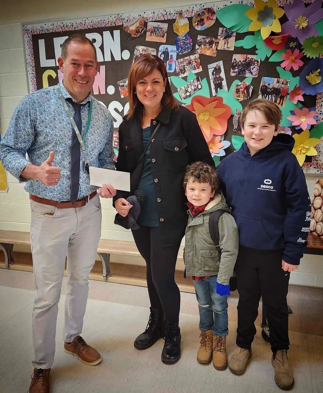 Four people posing indoors in front of a colorful bulletin board, smiling and holding donation cheque