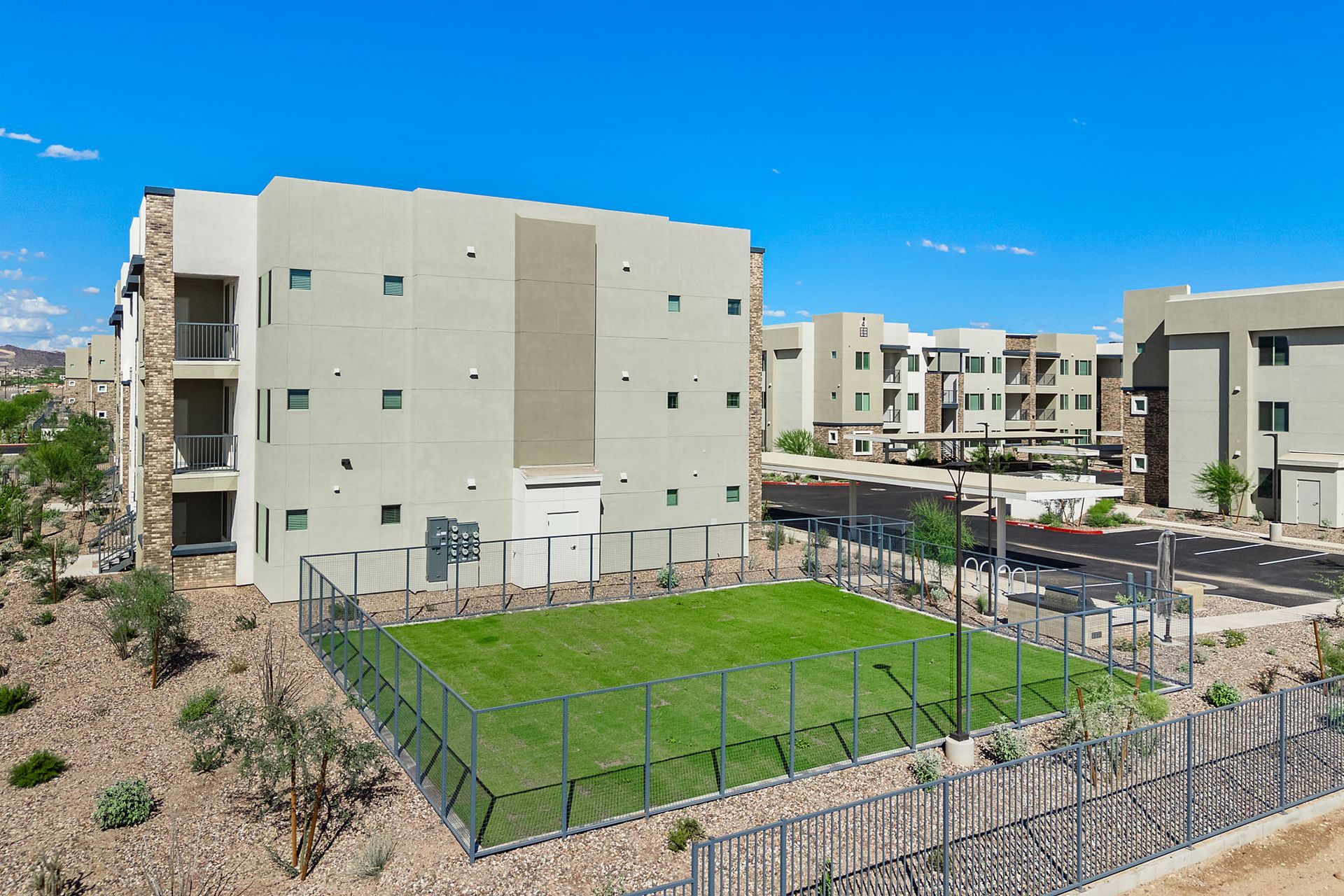 Apartment complex with a small fenced-in grassy area at Marq Bronco Butte, offers  1, 2, and 3 bedroom apartments in North Valley, Phoenix. Tan buildings with balconies, against a bright blue sky.