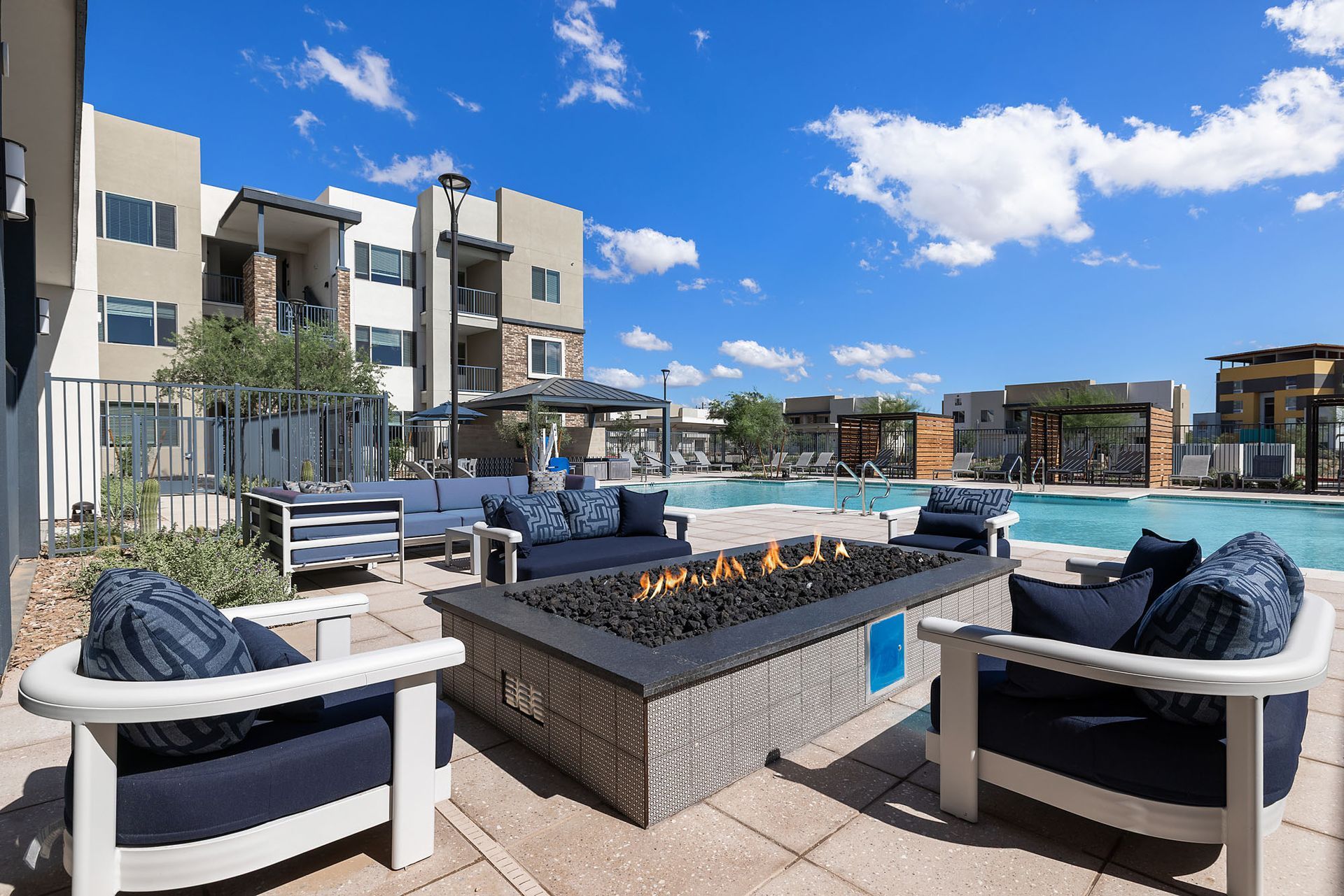 Outdoor lounge area with a fire pit, couches, and a swimming pool under a blue sky at Marq Bronco Butte, offers apartments near Dove Valley Towne Center in North Phoenix, AZ.