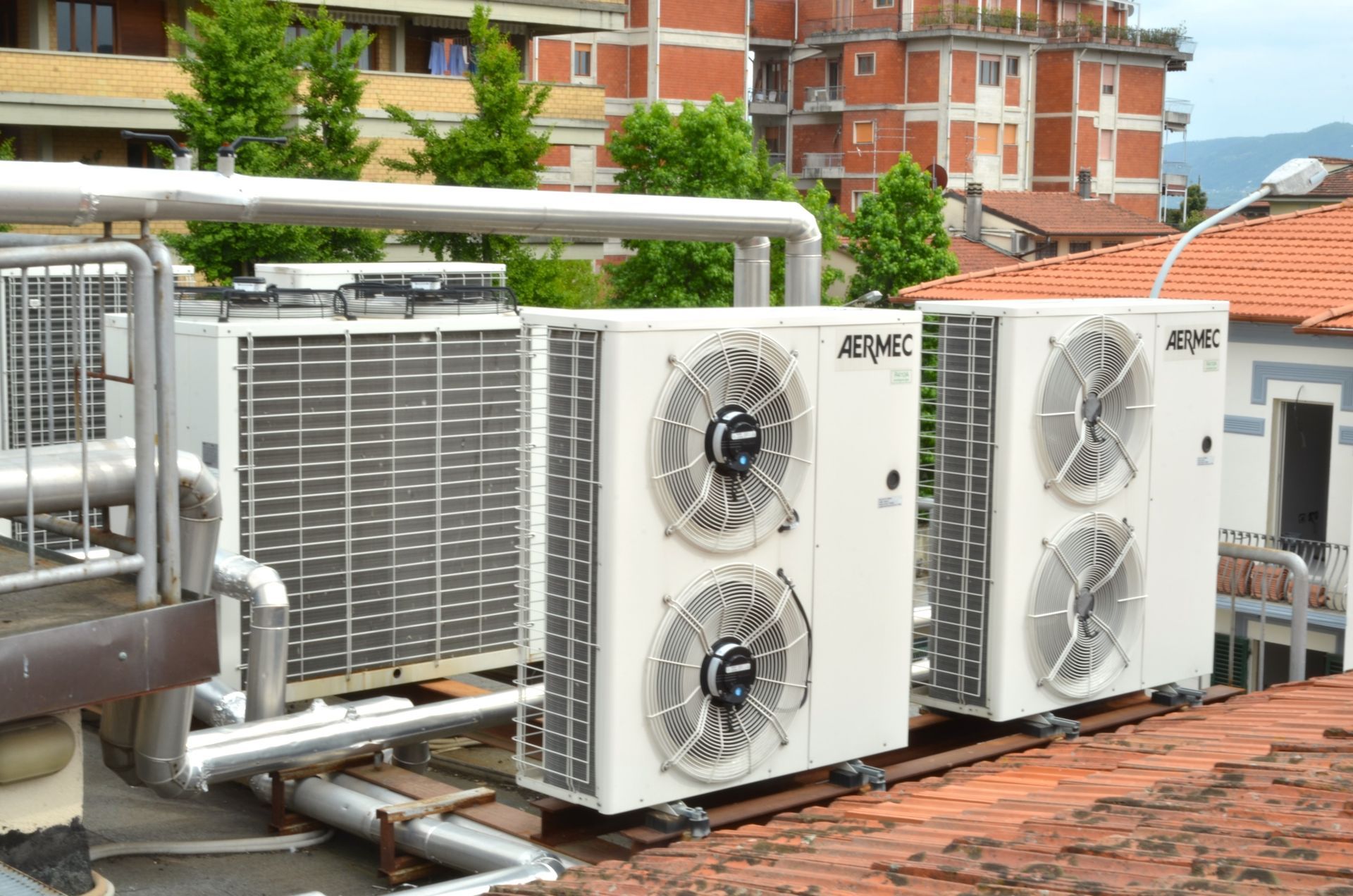 Unità esterne di climatizzazione e canalizzazioni su un tetto con edifici in mattoni sullo sfondo.