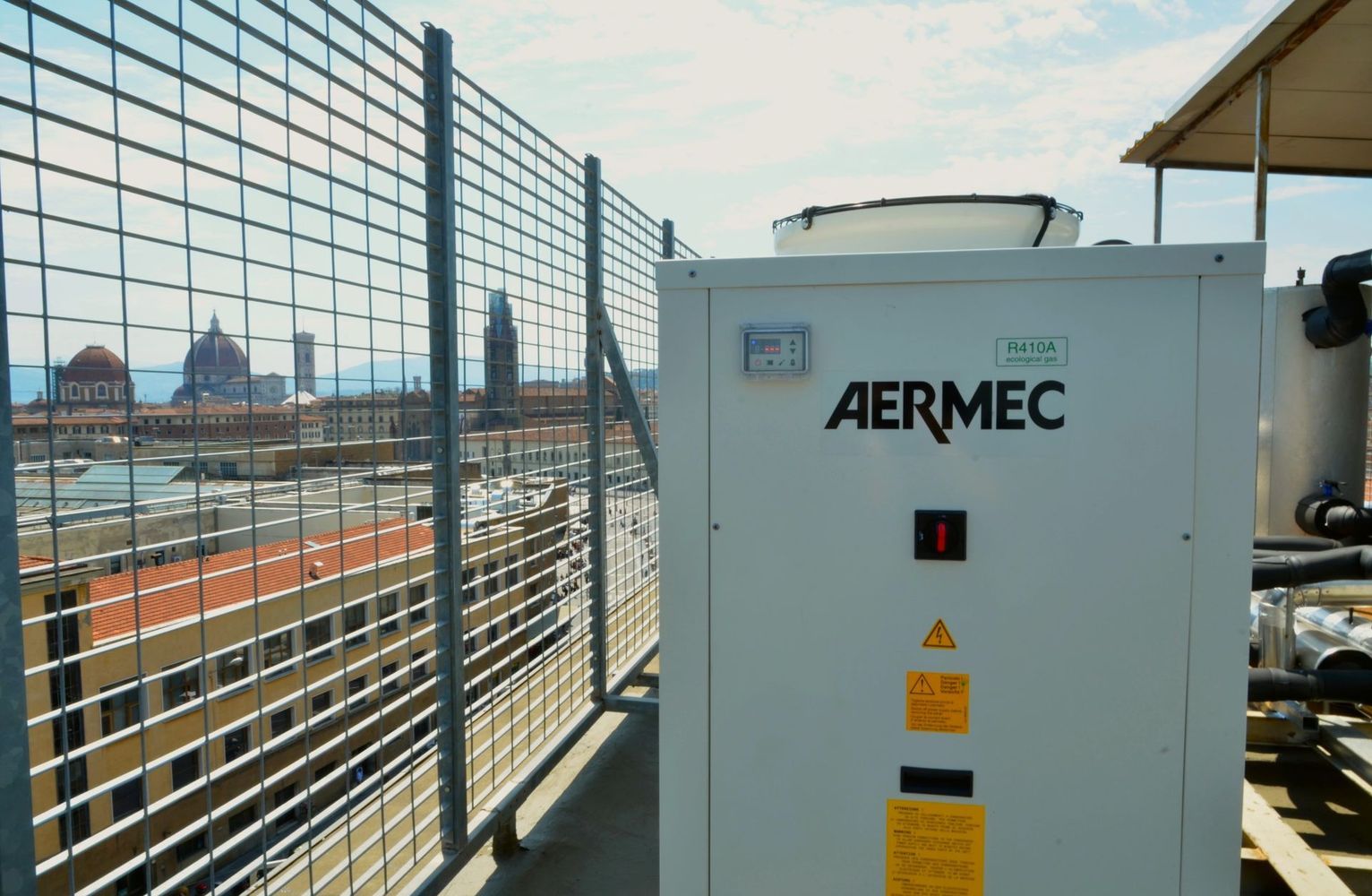 Unità di climatizzazione da tetto AERMEC bianca accanto a una recinzione metallica su un tetto industriale sotto un cielo luminoso