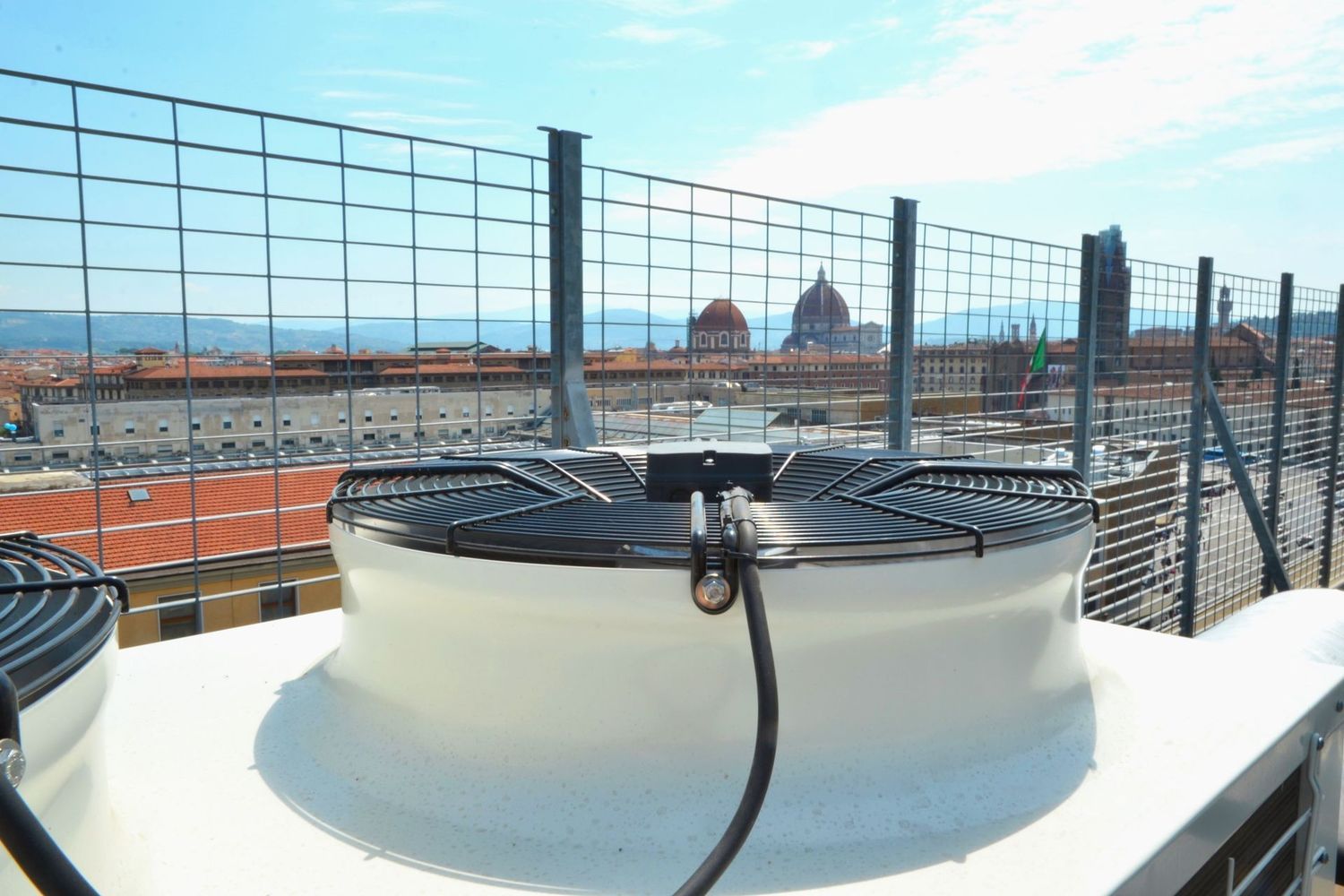 Unità di climatizzazione bianca sul tetto con ventilatore dietro una recinzione di sicurezza metallica sotto un cielo azzurro.