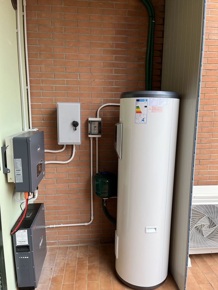 Nicchia di servizio con scaldabagno bianco, scatole elettriche e tubature a vista su una parete di mattoni.