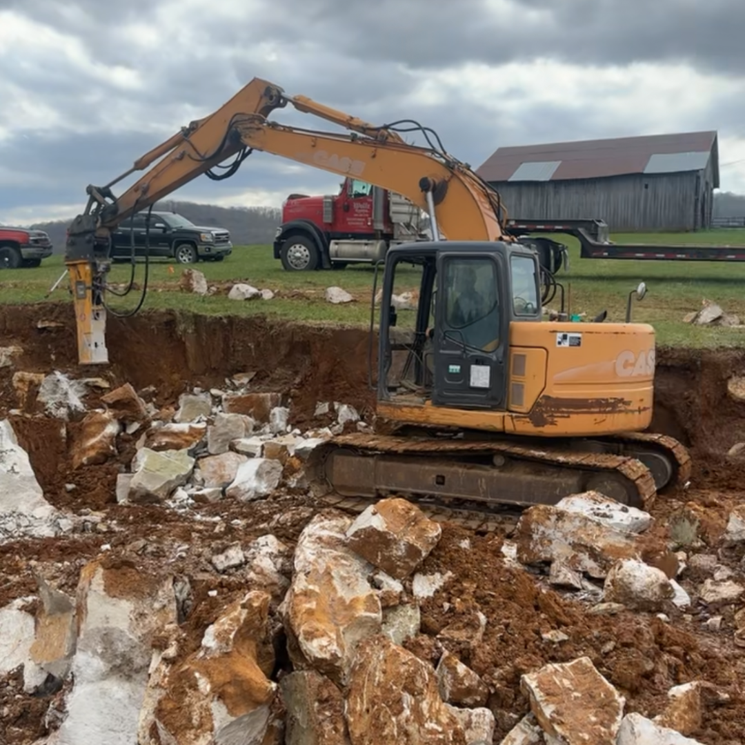 Excavator demolishing concrete in a dirt pit on a farm. A red truck and barn are in the background.