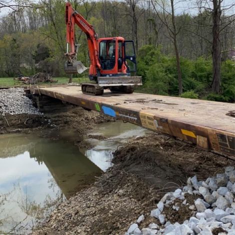 An excavator on a flatbed railcar bridge crossing a muddy creek.