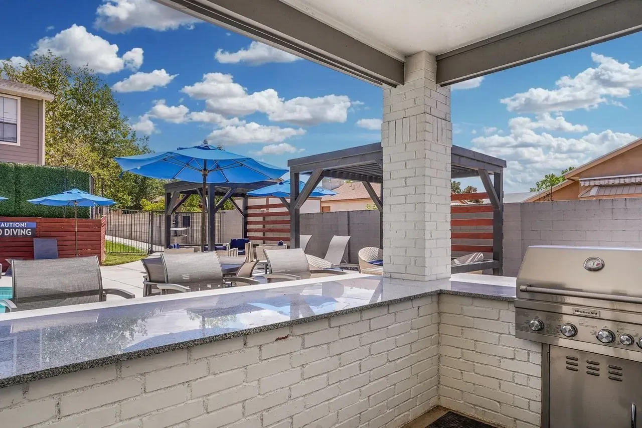 Outdoor kitchen and seating area with grills, umbrellas, and lounge chairs near a pool.
