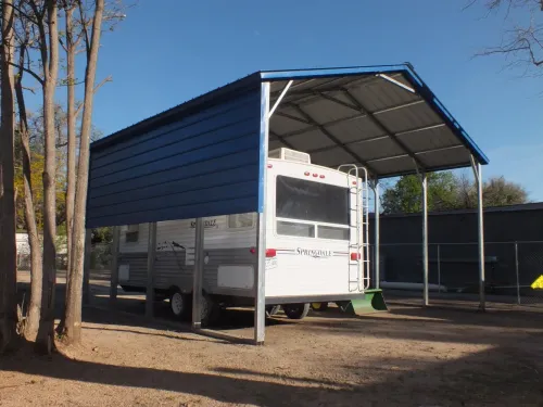 White RV parked under a blue metal cover on a dirt lot, with trees and a fenced area nearby